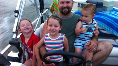Dad driving boat with three children on his lap.