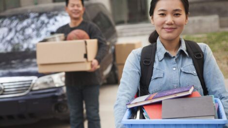 Asian college student carrying box of books with man in the background.
