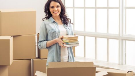 Young woman holding a stack of books surrounded by moving boxes.