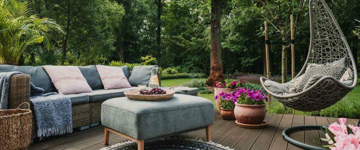 A patio garden is set up in a wooded area, with blue furniture and pink flowers set out on a stained deck.