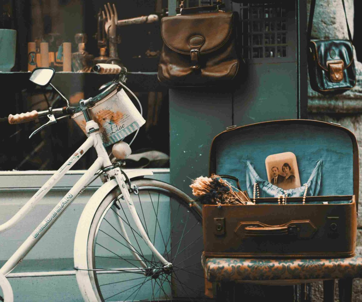 A vintage bicycle with a basket, a suitcase with a personal photo, dried flowers and jewelry along with personal bags hanging near a shop of antiques.