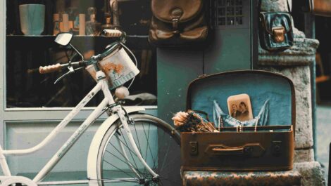 A vintage bicycle with a basket, a suitcase with a personal photo, dried flowers and jewelry along with personal bags hanging near a shop of antiques.